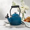 A Le Creuset Demi Kettle in Marseille sits on a marble countertop alongside muffins, a kitchen towel, and flowers in a vase.