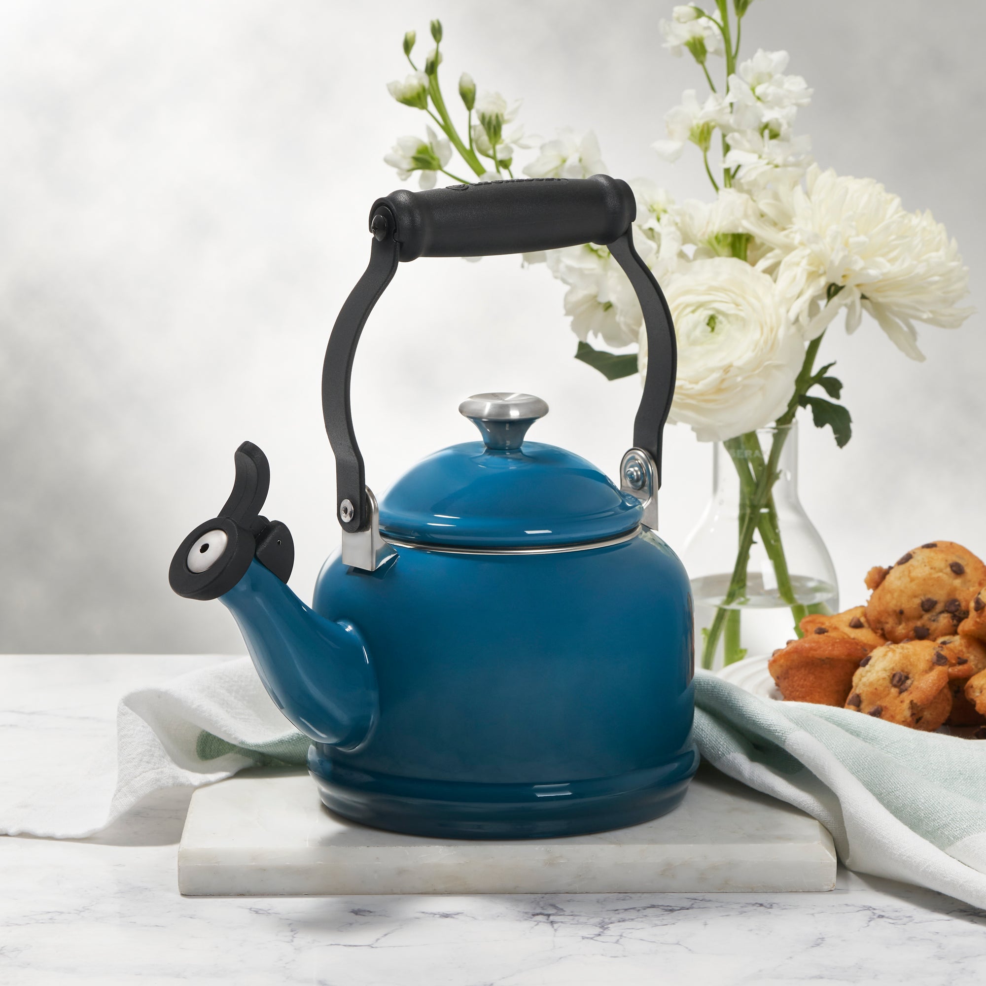 A Le Creuset Demi Kettle in Marseille sits on a marble countertop alongside muffins, a kitchen towel, and flowers in a vase.