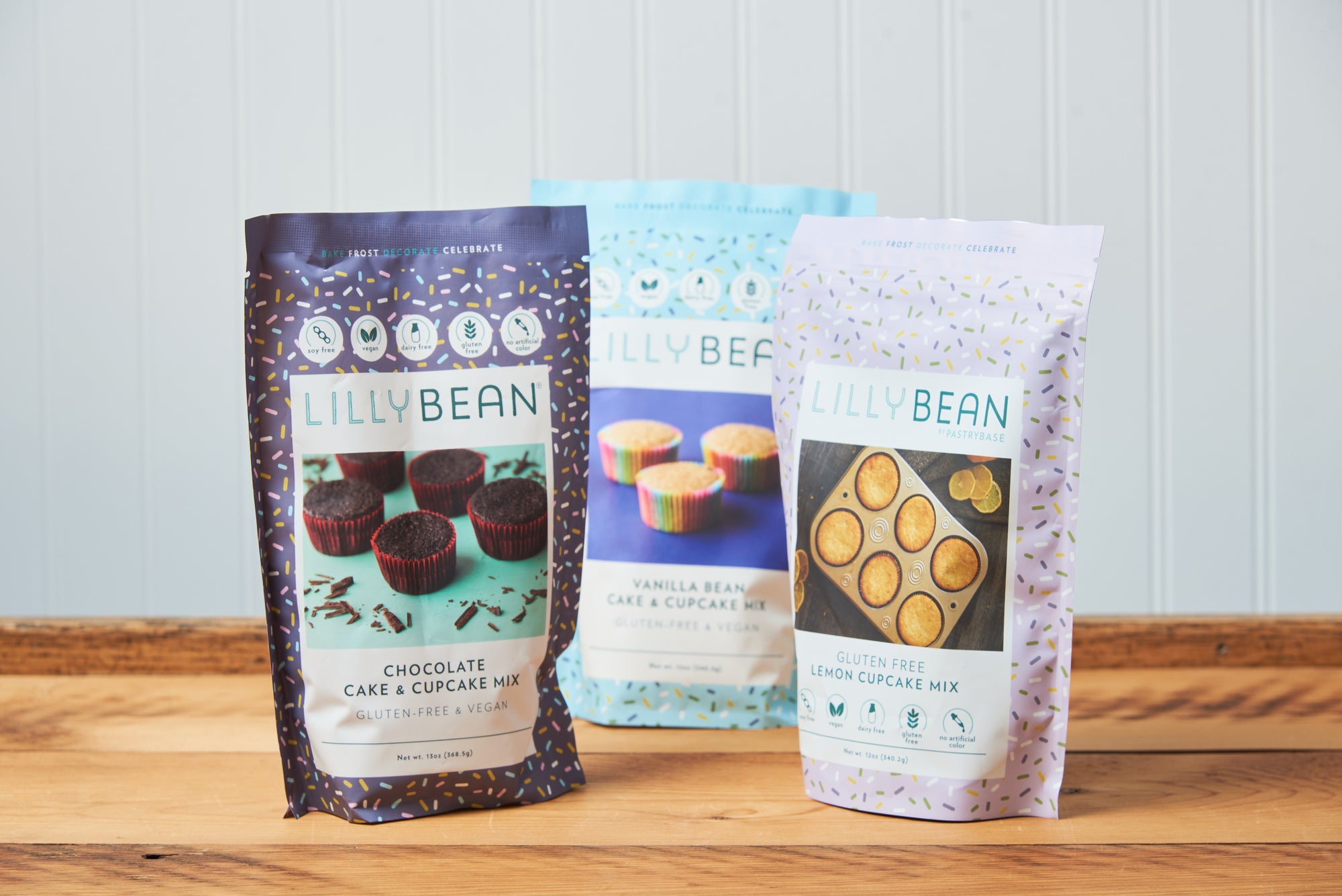 Three bags of LillyBean Cupcake Mix sit on a wooden tabletop. The flavors are Chocolate, Lemon, and Vanilla Bean.