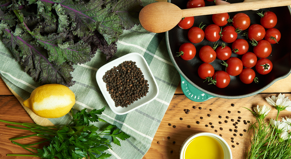 A green skillet full of tomatoes and garlic sits on a brown, wooden table surrounded by leafy greens and peppercorns.