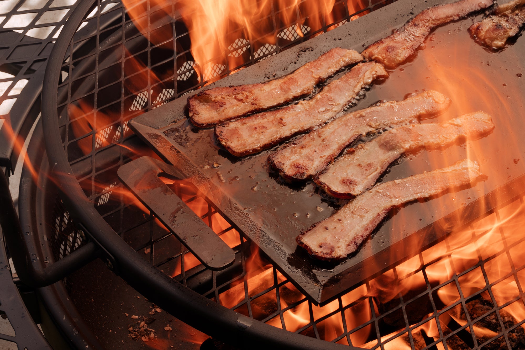 Bacon is cooked in a firepit on a Made In Seasoned Carbon Steel Griddle.