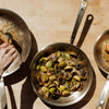 Three Made In Stainless Clad Frying Pans sits on wooden tabletop. Each fry pan contains food, including a steak, Brussel sprouts, and scallops. 