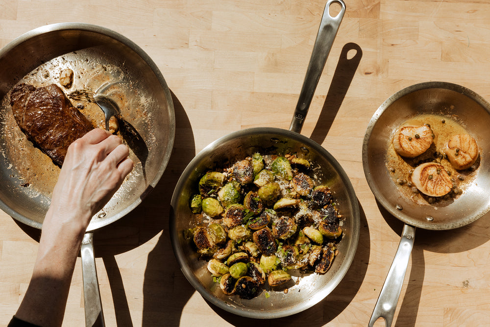 Three Made In Stainless Clad Frying Pans sits on wooden tabletop. Each fry pan contains food, including a steak, Brussel sprouts, and scallops. 