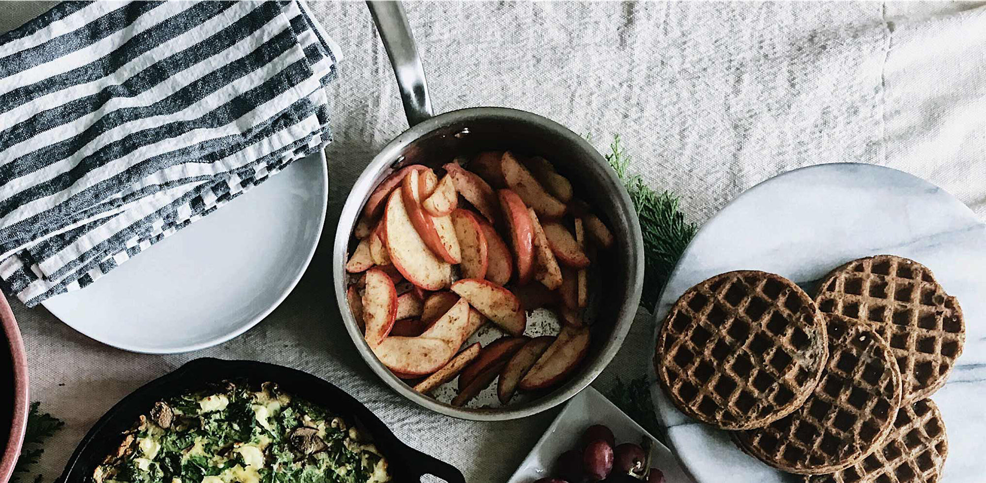 A Made In Stainless Clad Saucepan full of spiced apples sits on a tablespread. Alongside the saucepan are other dishes, including a plate of waffles.
