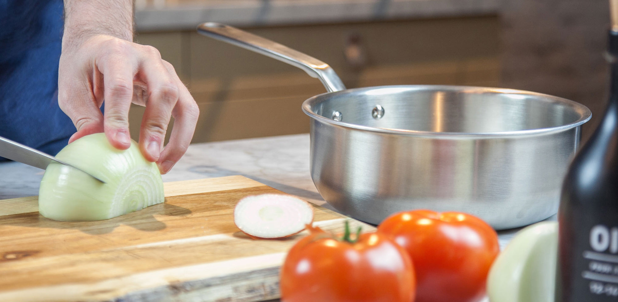 A Made In Stainless Clad Saucier sits on a countertop. Alongside it, a person cuts an onion.