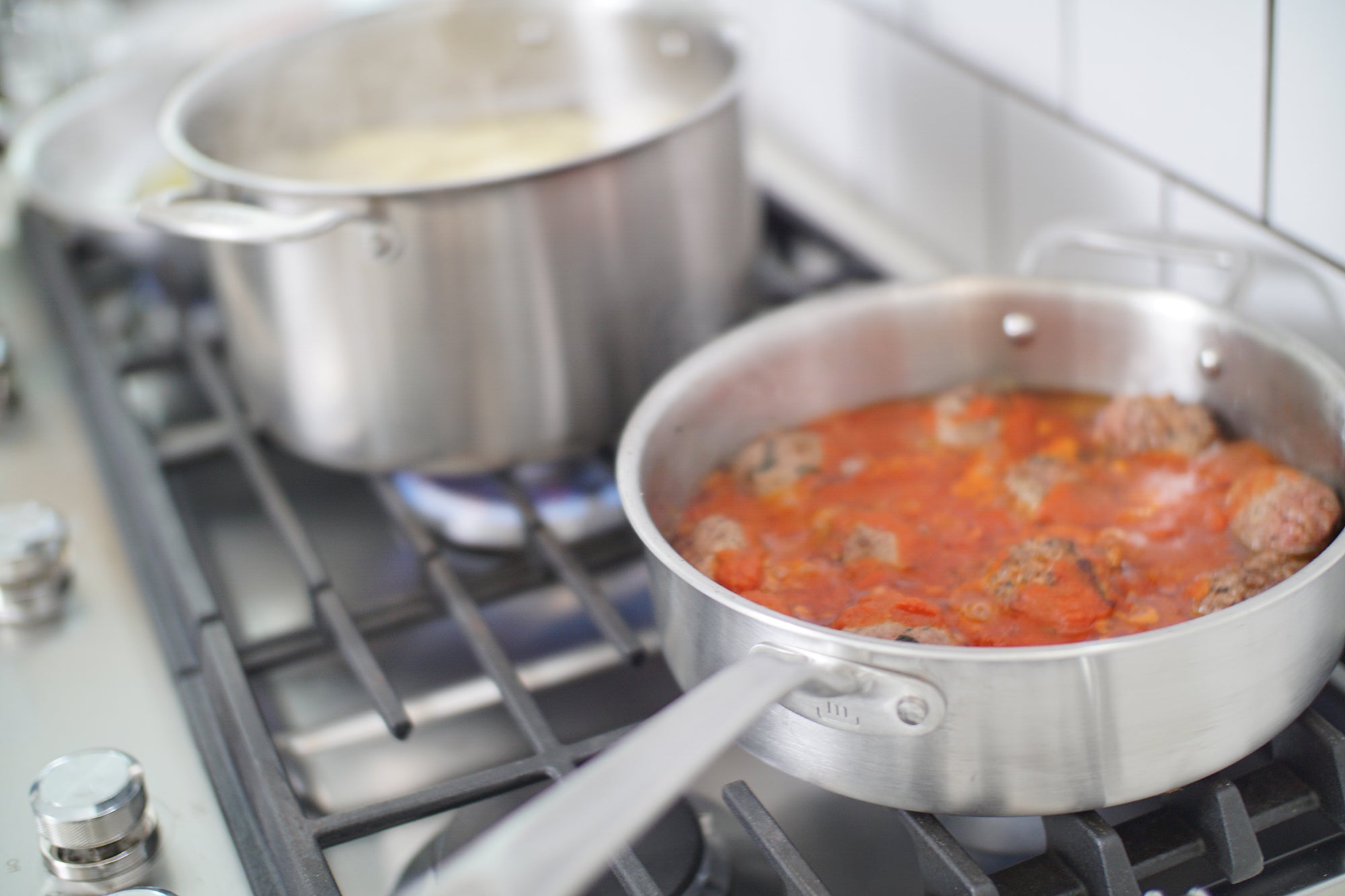 A Made In Stainless Clad Saute Pan on a stove top alongside a pot of noodles in boiling water. Meatballs and sauce are being cooked in the saute pan.