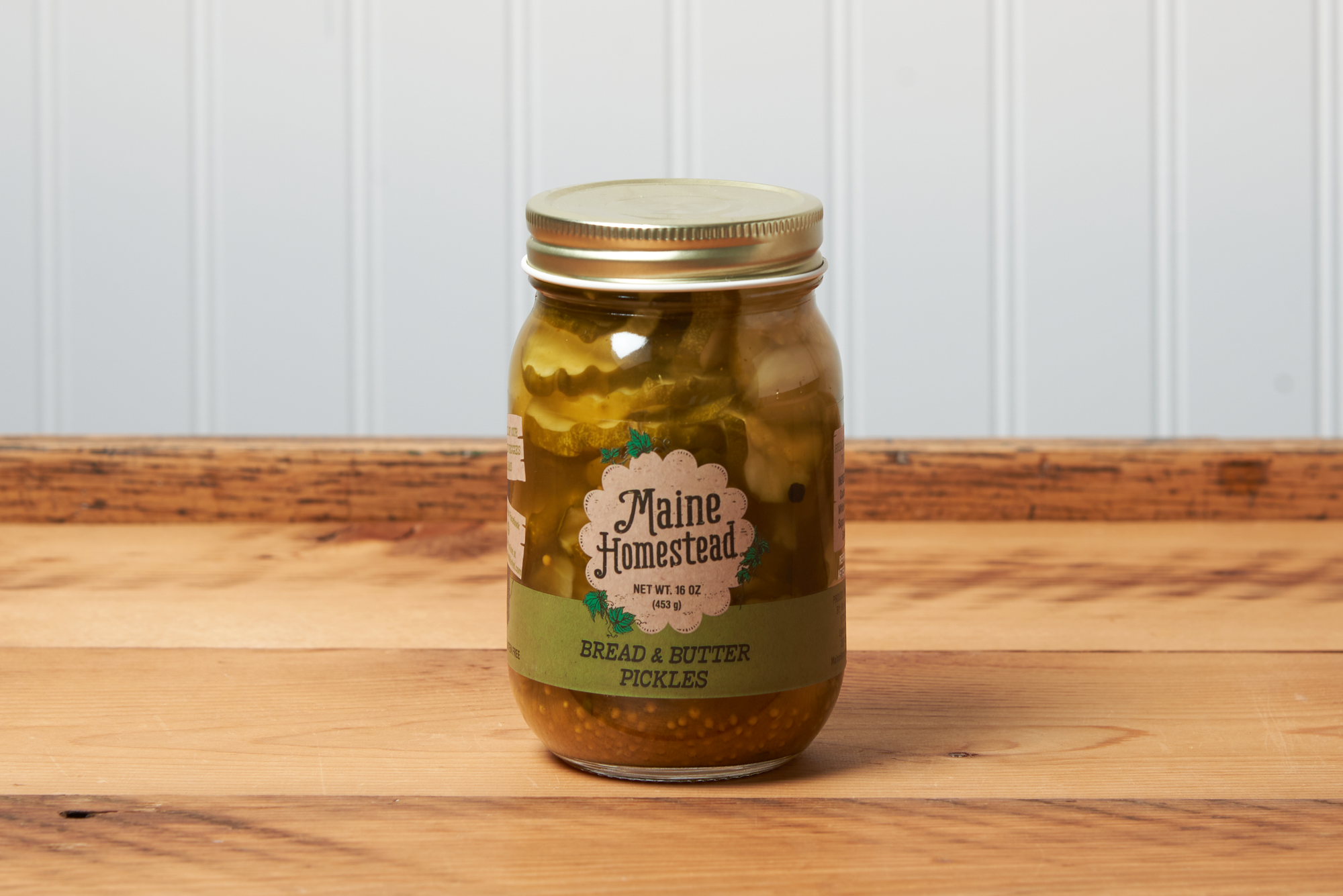 A jar of Maine Homestead Bread and Butter Pickles sits on a brown wooden table.