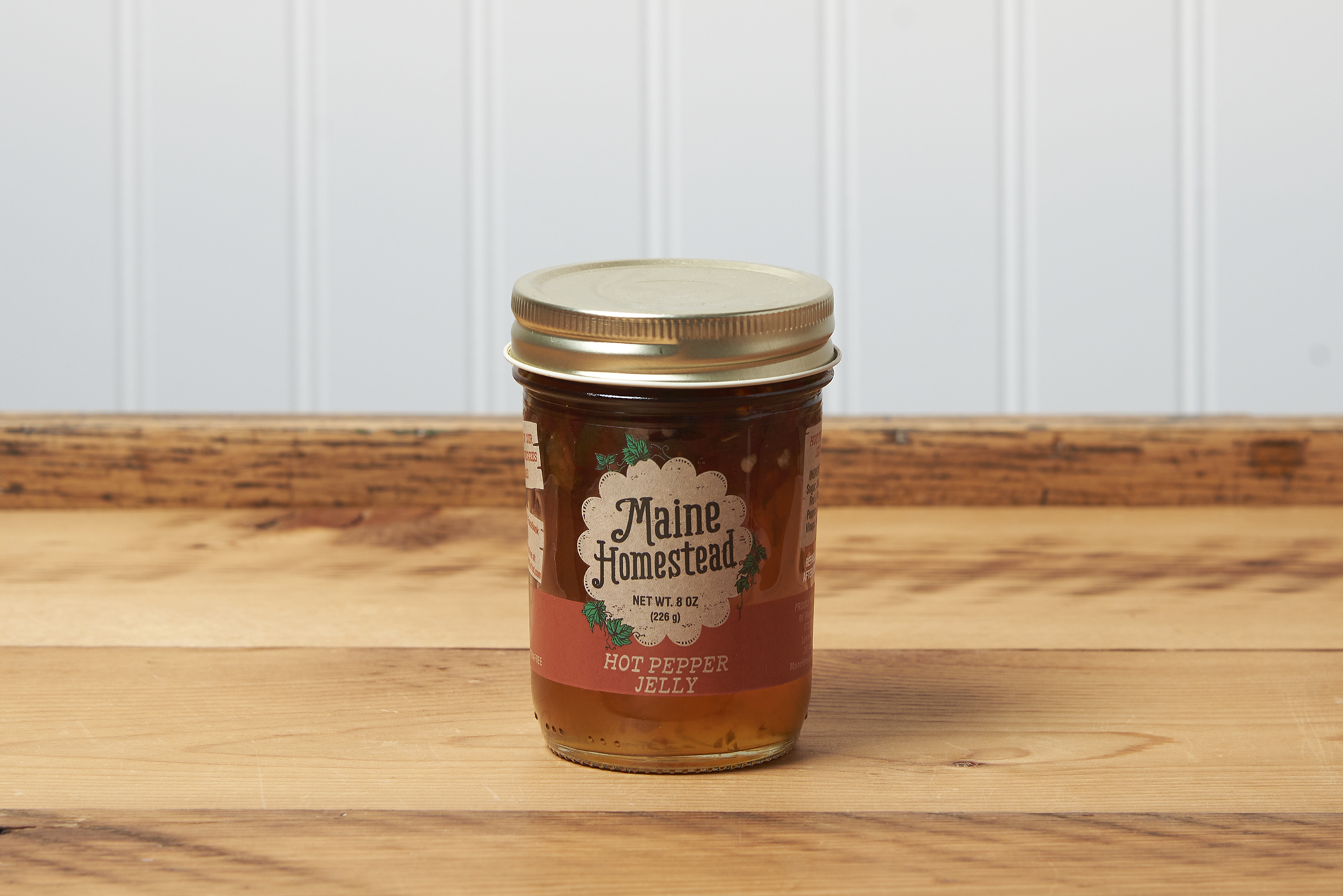 A jar of Maine Homestead Hot Pepper Jelly sits on a brown wooden table.