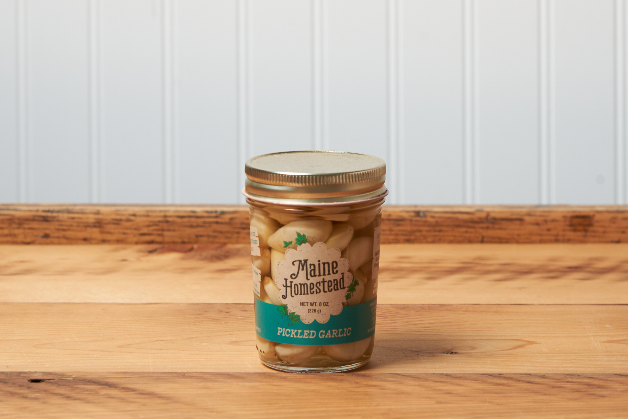 A jar of Maine Homestead Pickled Garlic sits on a brown wooden table.