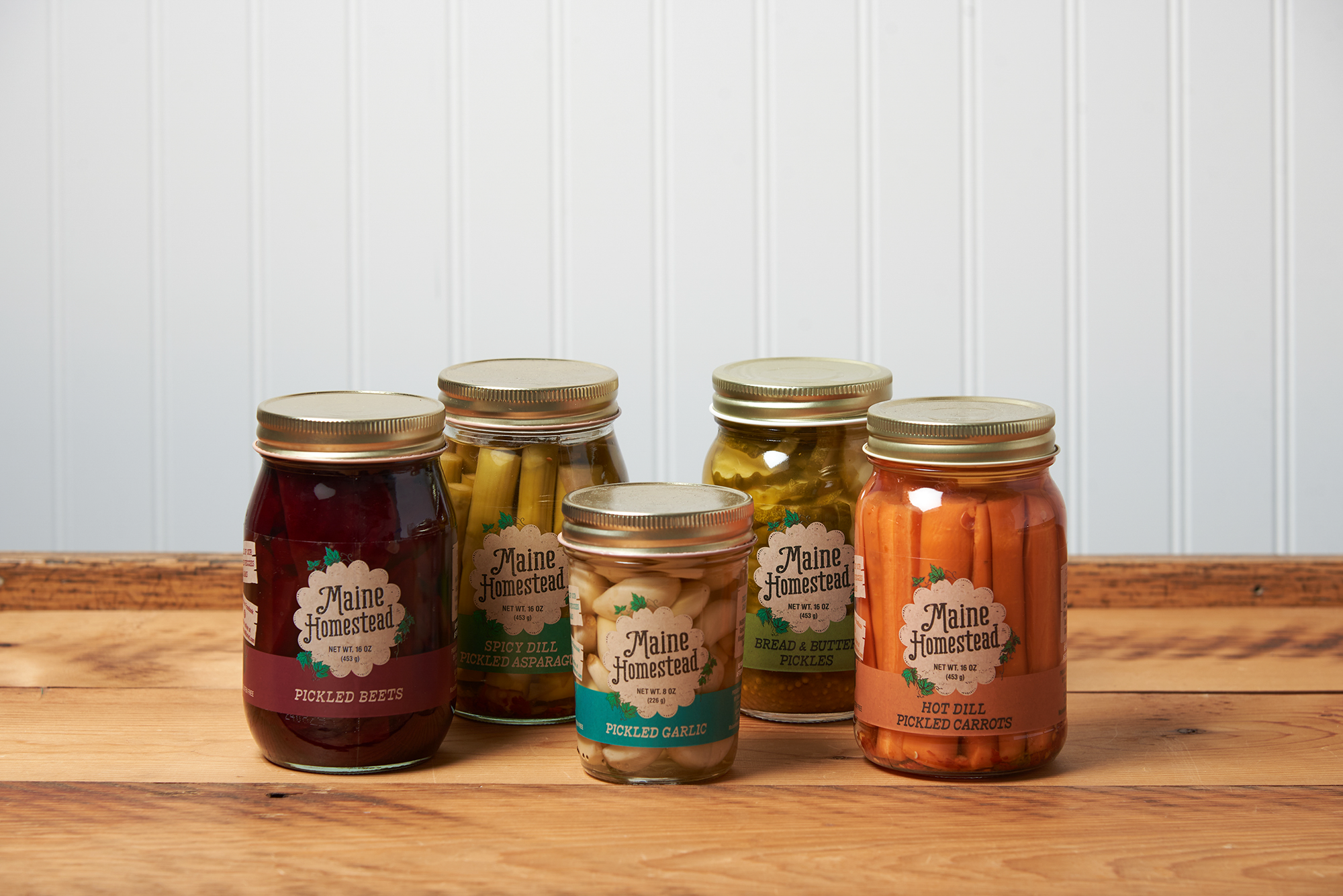 Five jars of assorted Maine Homestead pickled vegetables sit on a brown wooden table.