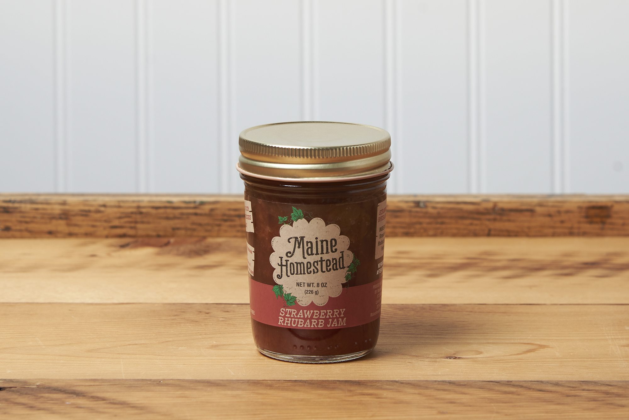A jar of Maine Homestead Strawberry Rhubarb Jam sits on a brown wooden table.