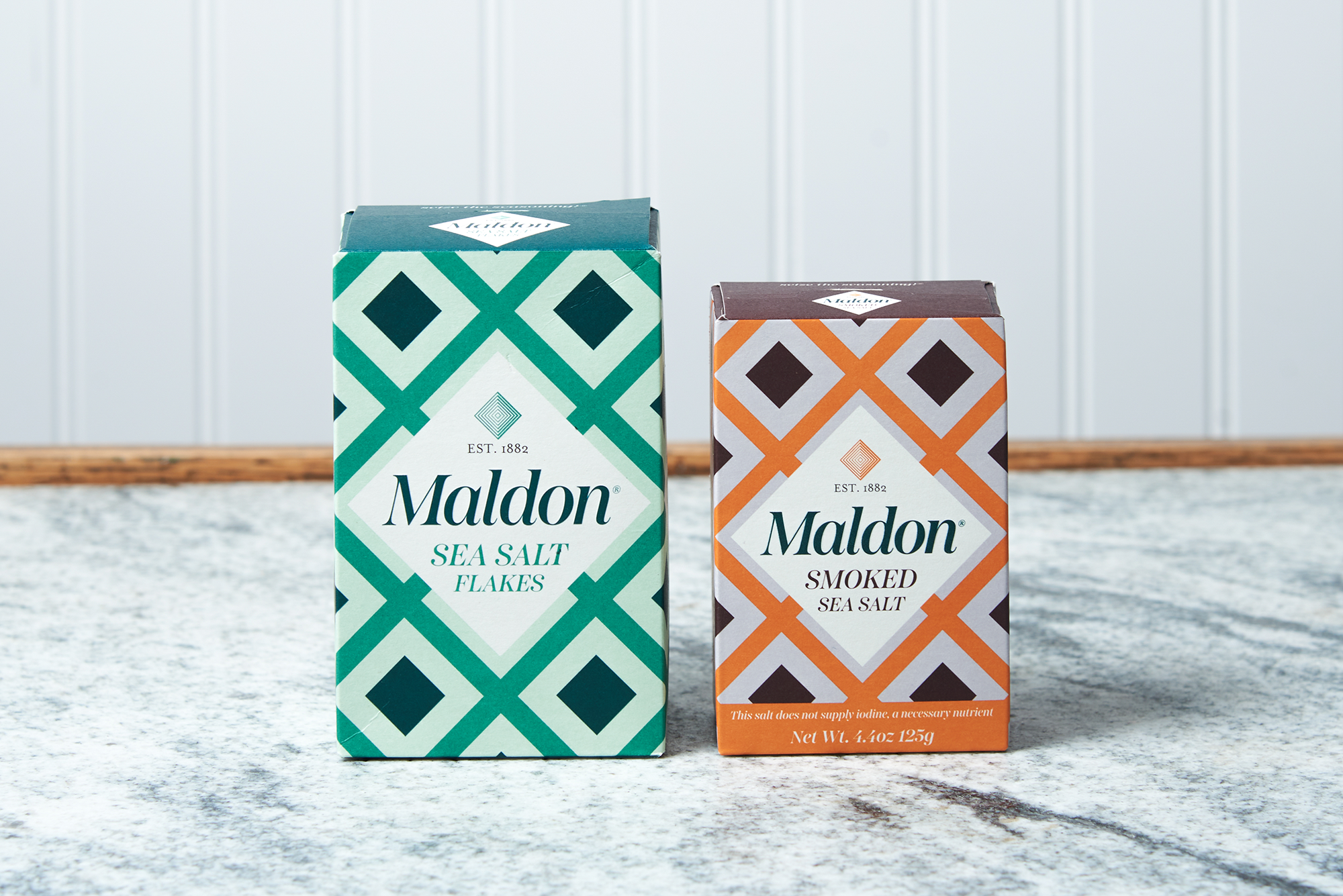 Two boxes of Maldon Sea Salt Flakes sit on a marble countertop. One box is classic Sea Salt Flakes, the other box is Smoked Sea Salt.