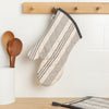 A Now Designs Pencil Stripe Grey Oven Mitt hangs on a kitchen wall.