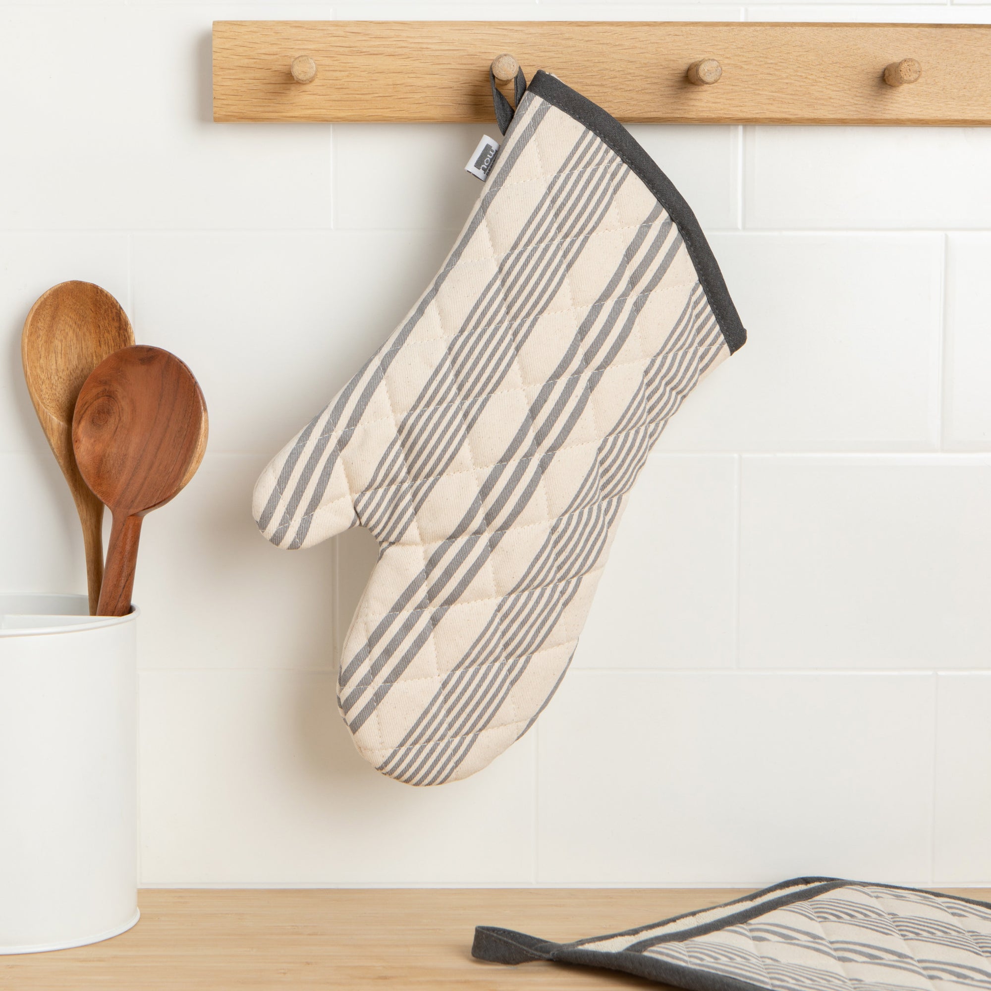 A Now Designs Pencil Stripe Grey Oven Mitt hangs on a kitchen wall.