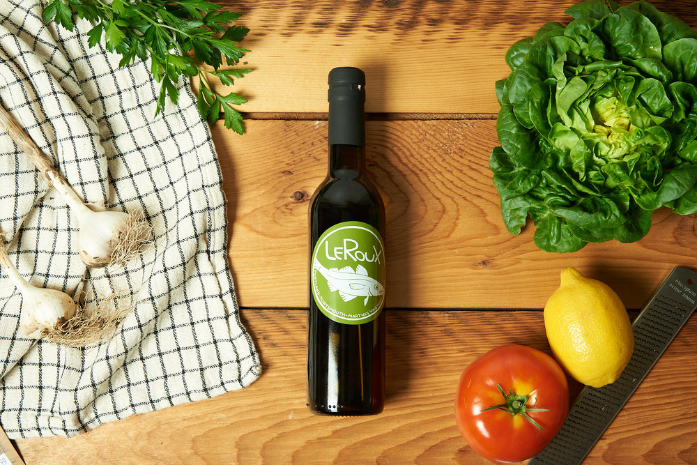 A bottle of olive oil sits on a wooden table surrounded by produce.