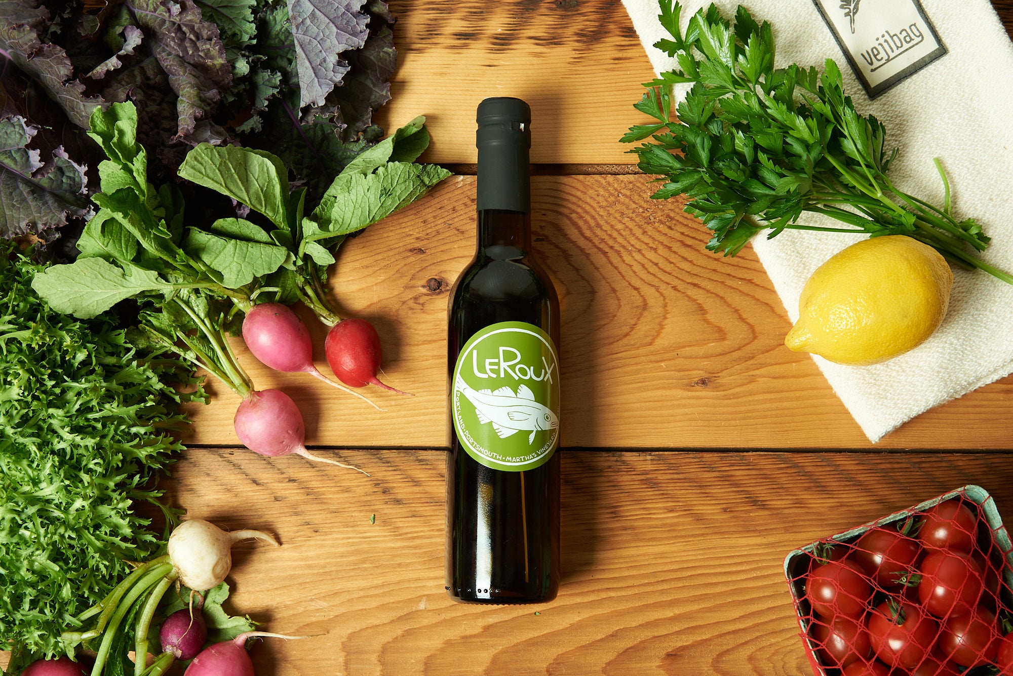 A bottle of olive oil sits on a wooden table surrounded by produce.