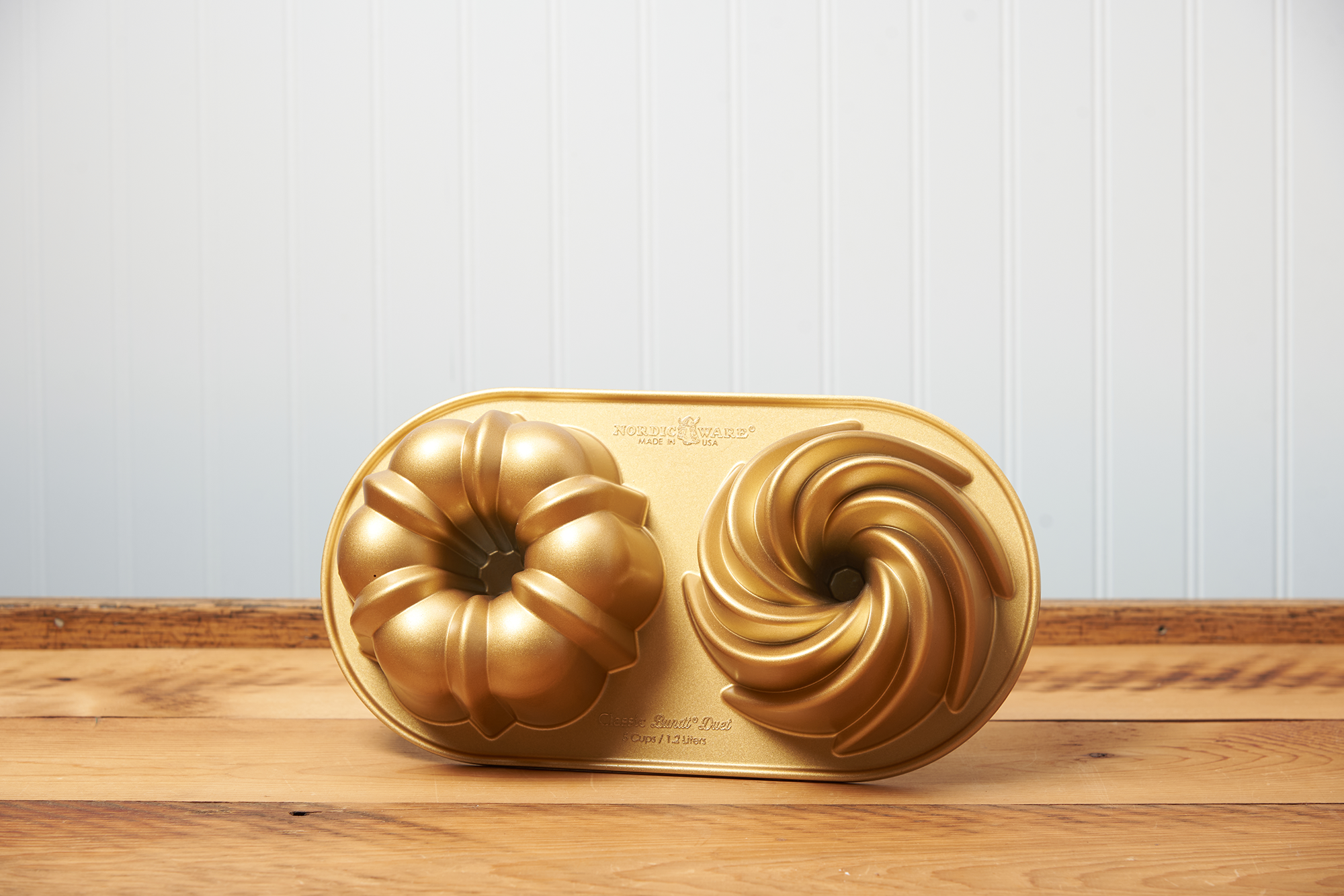 A Nordic Ware Classic Bundt Duet Pan sits on a brown, wooden table.