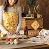 A woman makes pasta on a wooden countertop. She wears a Now Designs Free Range apron.