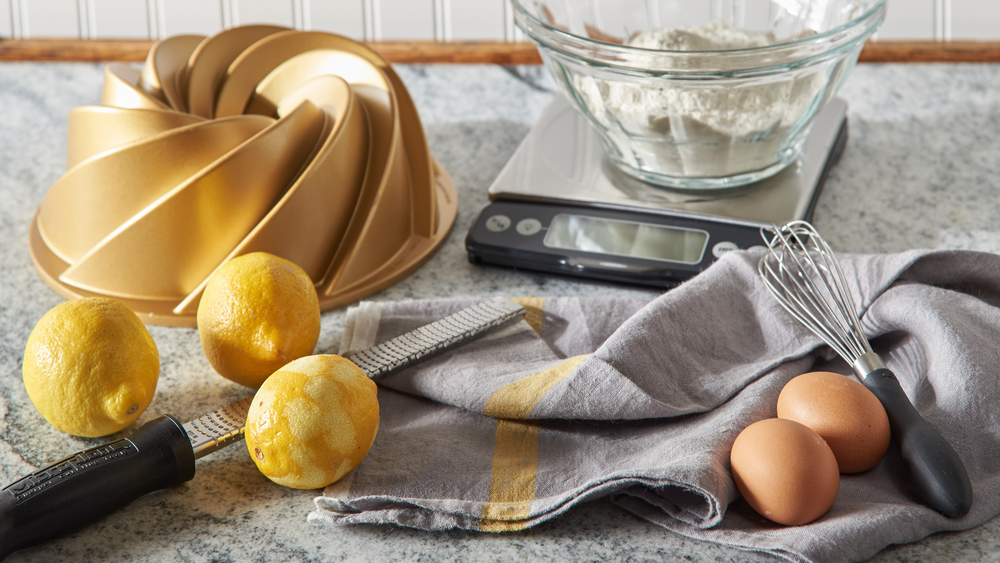 Kitchen scene with bundt cake pan, lemons, eggs, whisk, and digital scale on countertop.