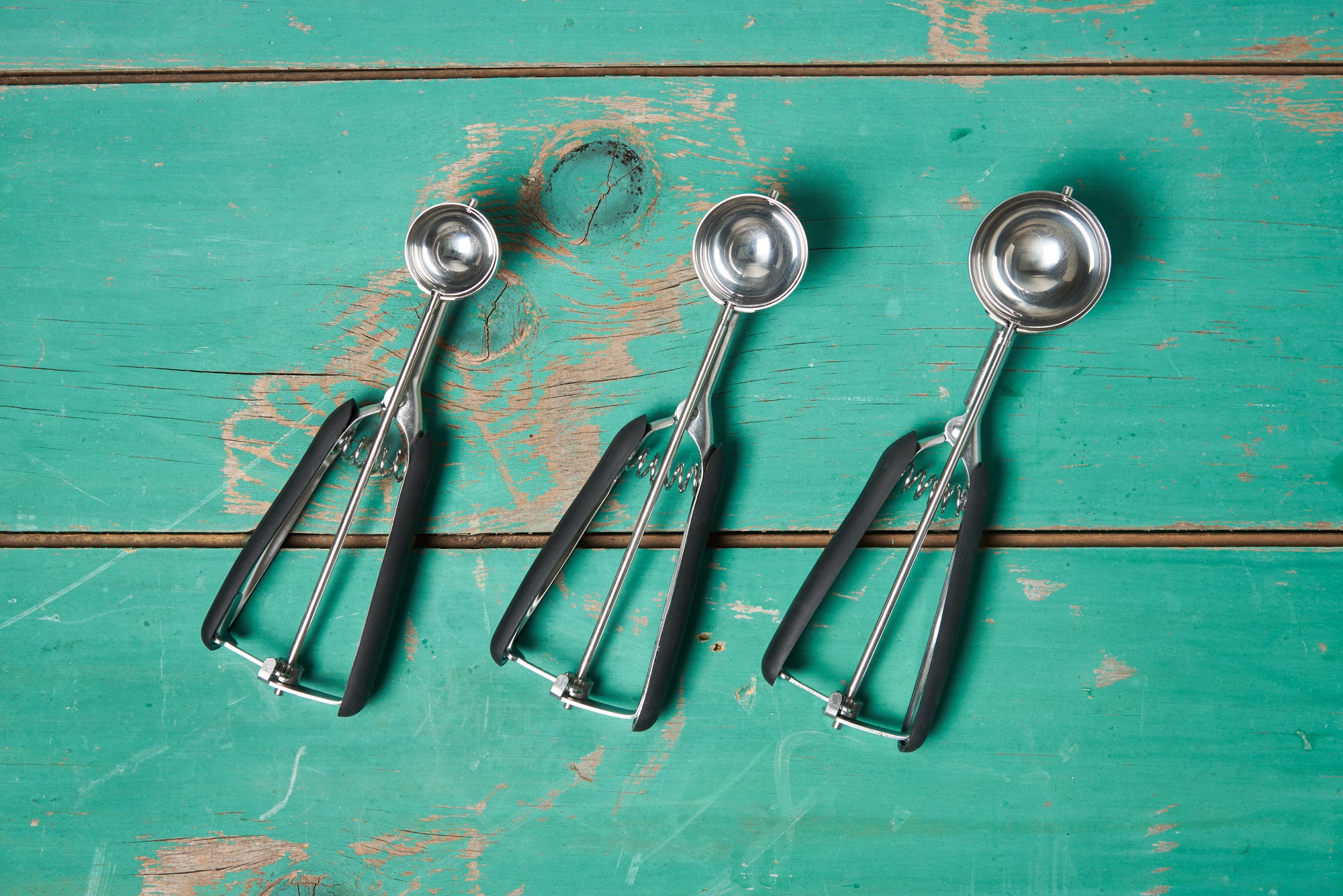 Three sizes of OXO Cookie Scoops— small, medium, and large— sit on a turquoise wooden tabletop.