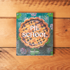 The book 'Pie School' by Kate Lebo is displayed flat on a rustic wooden plank background. The book cover features a detailed illustration of a lattice-pie crust in the center, set against a dark teal background decorated with floral sketches and white title text.