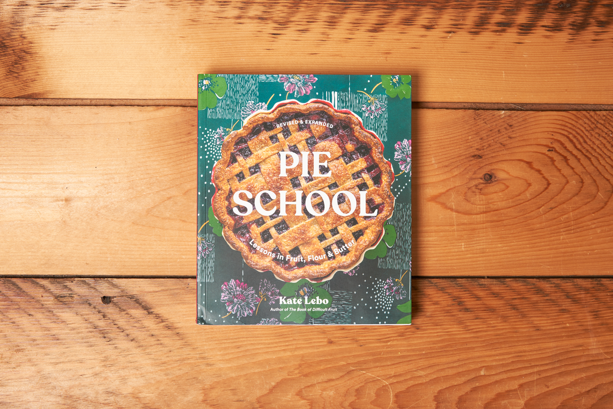 The book 'Pie School' by Kate Lebo is displayed flat on a rustic wooden plank background. The book cover features a detailed illustration of a lattice-pie crust in the center, set against a dark teal background decorated with floral sketches and white title text.