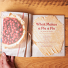 The book 'Pie School: Lessons in Fruit, Flour & Butter' sits open on a wooden tabletop. A hand holds open a page with a photo of an in-progress cherry pie alongside the title "What Makes a Pie a Pie"