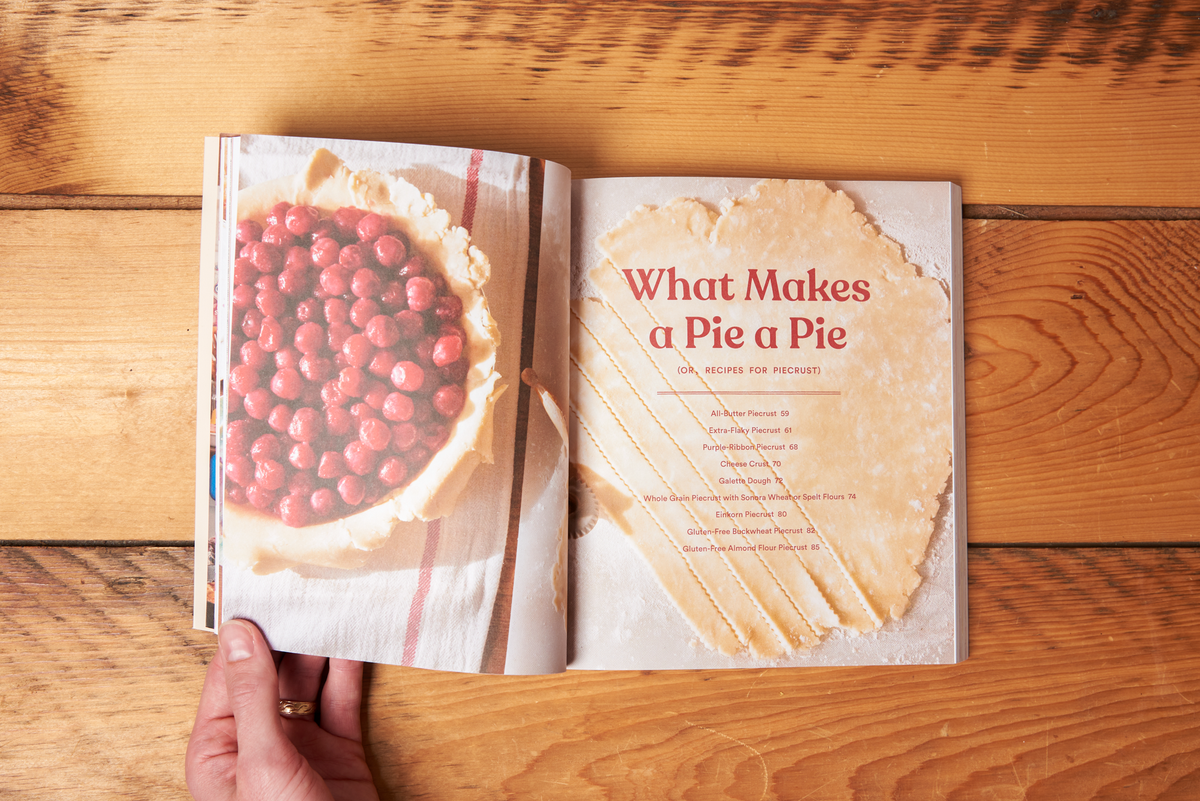 The book 'Pie School: Lessons in Fruit, Flour & Butter' sits open on a wooden tabletop. A hand holds open a page with a photo of an in-progress cherry pie alongside the title "What Makes a Pie a Pie"