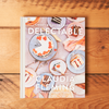 The cookbook 'Delectable: Sweet & Savory Baking' sits closed on a wooden tabletop. The cover features an assortment of baked desserts.