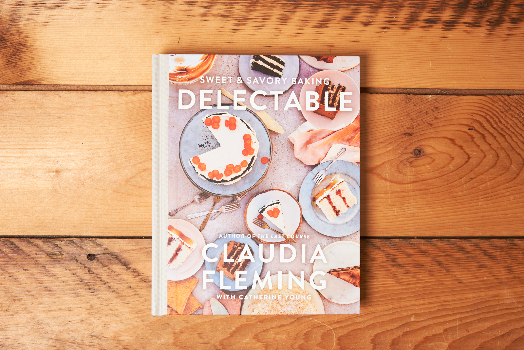 The cookbook 'Delectable: Sweet & Savory Baking' sits closed on a wooden tabletop. The cover features an assortment of baked desserts.