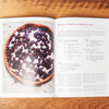 The book 'Delectable: Sweet & Savory Baking' sits open on a wooden tabletop. The open page shows a photo blueberry tart alongside its recipe, title "Blueberry, Blueberry, Blueberry Tart."