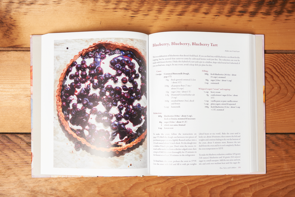 The book 'Delectable: Sweet & Savory Baking' sits open on a wooden tabletop. The open page shows a photo blueberry tart alongside its recipe, title "Blueberry, Blueberry, Blueberry Tart."