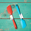 A red and turquoise Ela's Favorite Silicone Spoon sits on a turquoise tabletop.