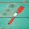 A red Ela's Favorite Silicone Spoon sits on a turquoise tabletop.
