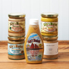 An assortment of Raye's Mustard products sit on a brown wooden table. The assortment includes the following mustards: Maple Horseradish, Brown Ginger, Maine Beer Company Lunch, Downeast Schooner, and Top Dog.