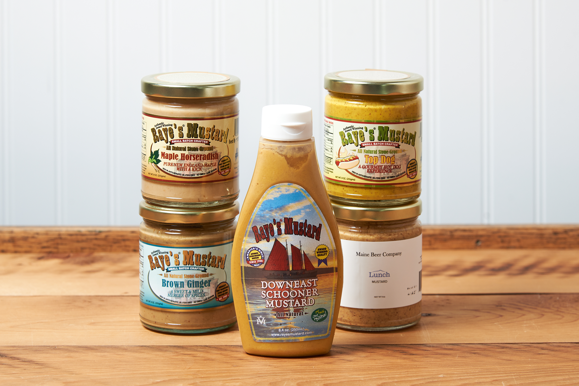 An assortment of Raye's Mustard products sit on a brown wooden table. The assortment includes the following mustards: Maple Horseradish, Brown Ginger, Maine Beer Company Lunch, Downeast Schooner, and Top Dog.