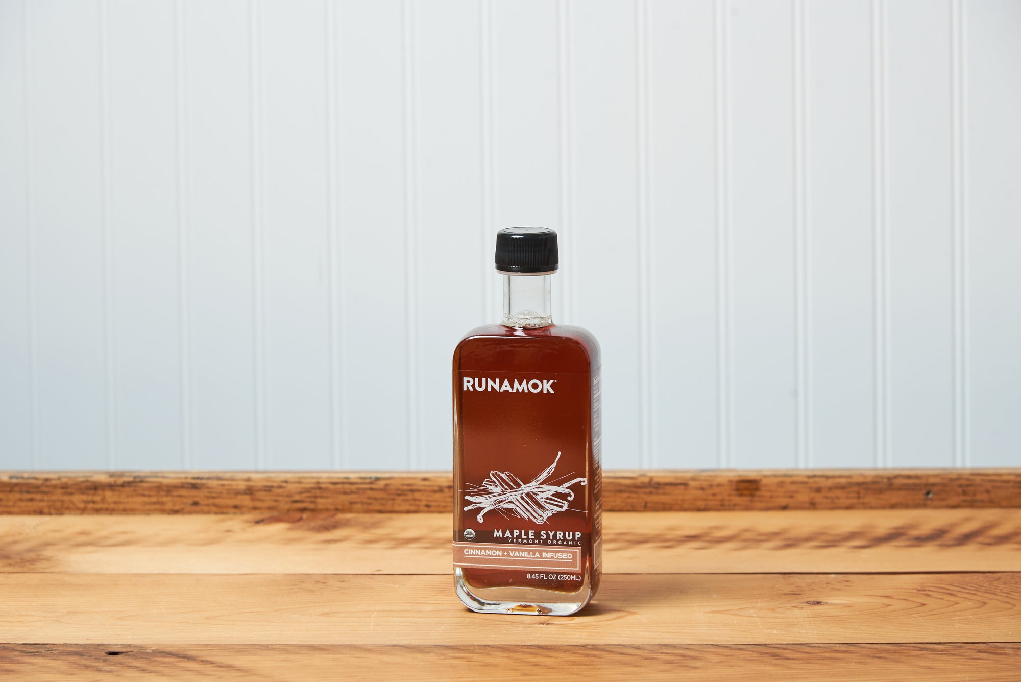 A bottle Runamok Cinnamon and Vanilla Infused Maple Syrup sits on a wooden tabletop.