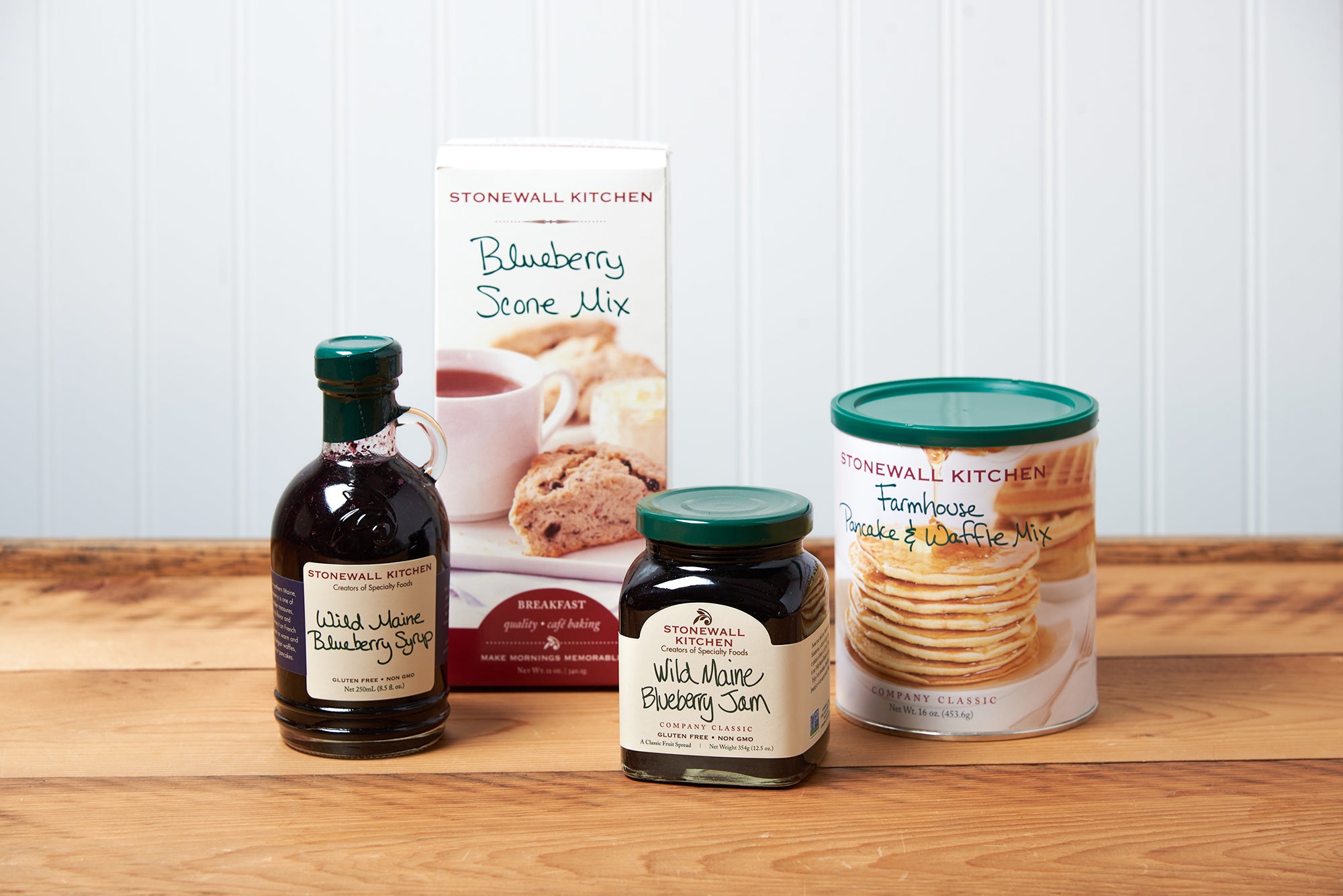 An assortment of Stonewall Kitchen items sit on a brown wooden table. The assortment includes: Wild Maine Blueberry Jam, Wild Maine Blueberry Syrup, Blueberry Scone Mix, and Farmhouse Pancake & Waffle Mix
