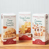 Three Stonewall Kitchen Baking Mixes on a wooden tabletop. The mixes include Orange Cranberry Scone, Cinnamon Bun, and Sugar Cookie.
