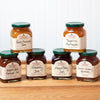 A group of six assorted Stonewall Kitchen jams and marmalades sit on a brown wooden table. The group includes the following flavors: Raspberry Peach Champagne Jam, Orange Cranberry Marmalade, Strawberry Jam, Mixed Berry Jam, Tangerine Marmalade, and Peach Amaretto Jam.