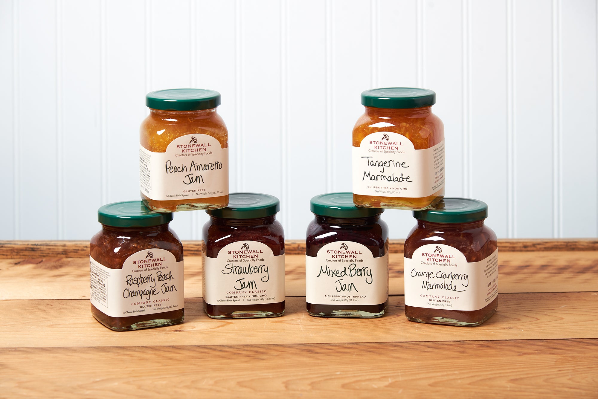 A group of six assorted Stonewall Kitchen jams and marmalades sit on a brown wooden table. The group includes the following flavors: Raspberry Peach Champagne Jam, Orange Cranberry Marmalade, Strawberry Jam, Mixed Berry Jam, Tangerine Marmalade, and Peach Amaretto Jam.