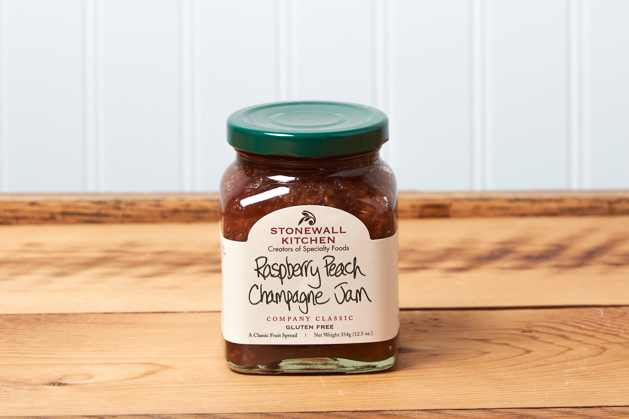 A jar of Stonewall Kitchen Raspberry Peach Champagne Jam sits on a brown wooden table.