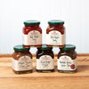 A group of five assorted Stonewall Kitchen jams and jellies sit on a brown wooden table.  The group includes the following flavors: Red Pepper Jelly, Hot Pepper Jelly, Roasted Garlic Onion Jam, Fig & Ginger Jam, and Maple Bacon Onion Jam.