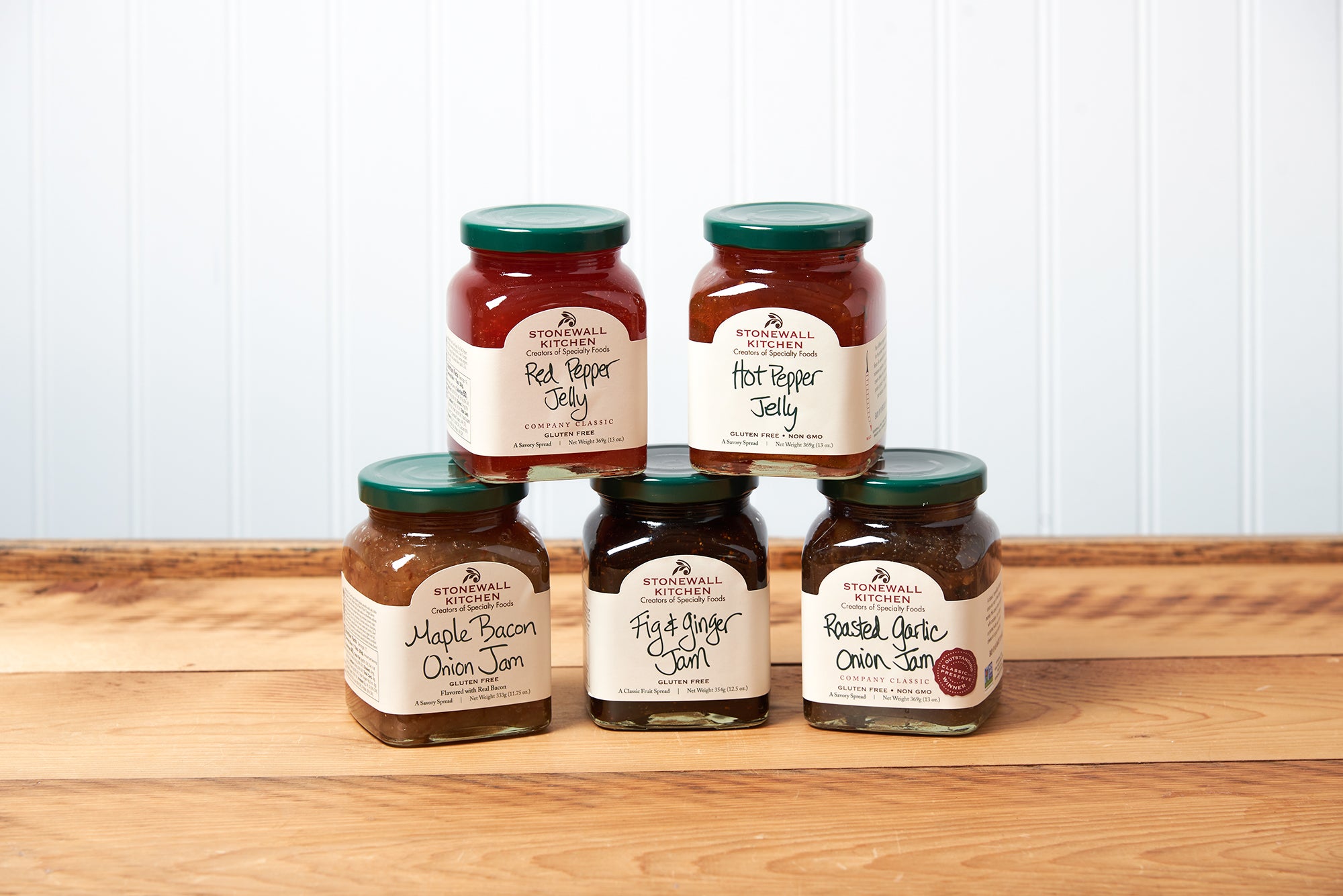 A group of five assorted Stonewall Kitchen jams and jellies sit on a brown wooden table.  The group includes the following flavors: Red Pepper Jelly, Hot Pepper Jelly, Roasted Garlic Onion Jam, Fig & Ginger Jam, and Maple Bacon Onion Jam.