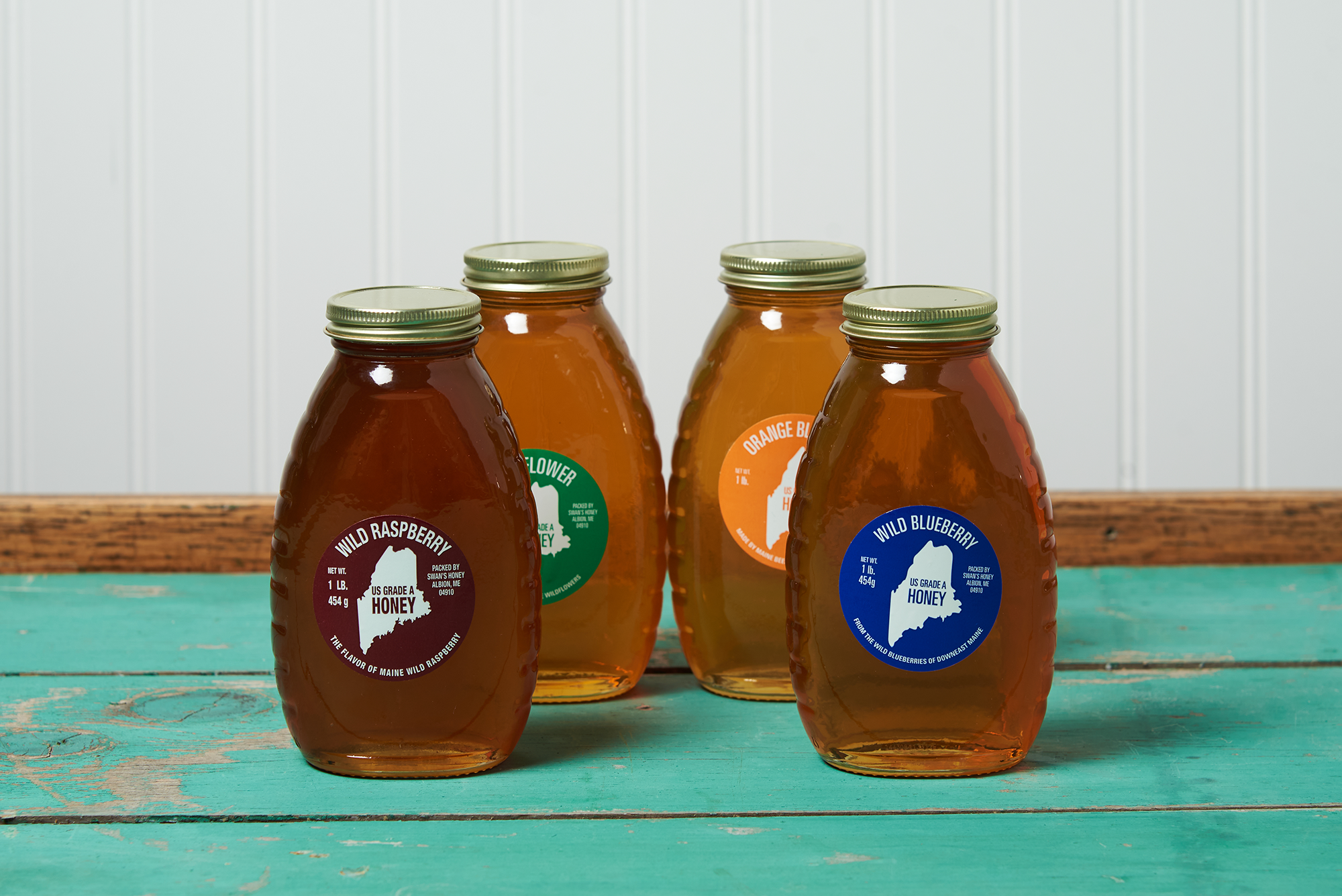 An assortment of four jars of Swan's Honey sits on a turquoise wooden table. the assortment includes the following honeys: Wild Raspberry, Wildflower, Orange Blossom, and Wild Blueberry
