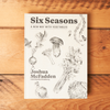 The cookbook 'Six Seasons: A New Way with Vegetables' sits closed on a wooden table. Illustrations of vegetables adorn the cover.