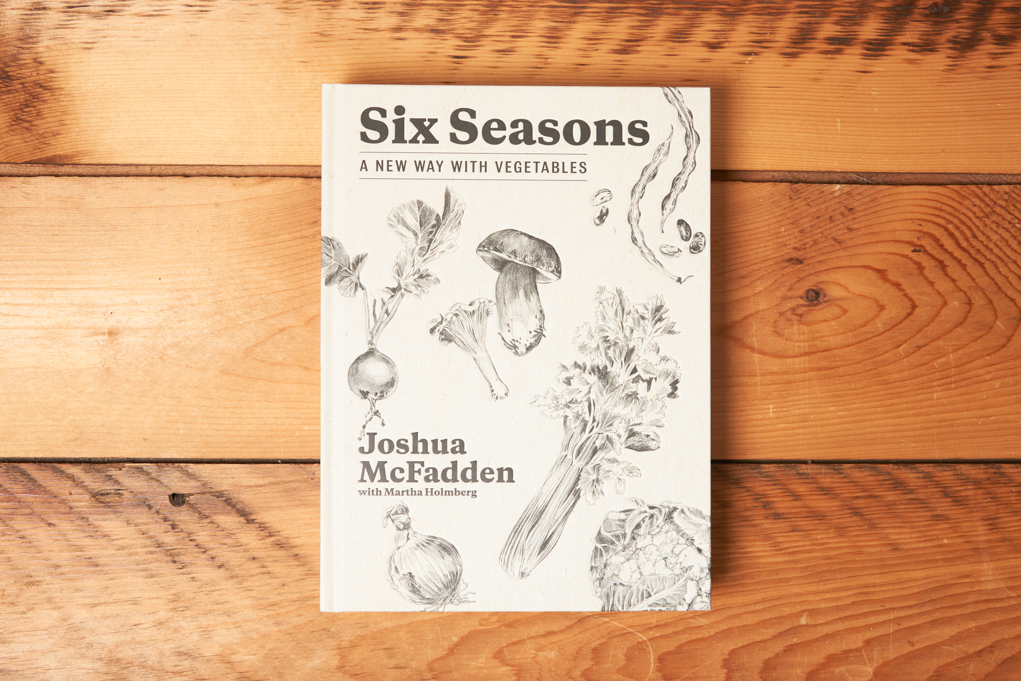 The cookbook 'Six Seasons: A New Way with Vegetables' sits closed on a wooden table. Illustrations of vegetables adorn the cover.