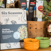 The cookbook 'Six Seasons: A New Way with Vegetables' sits on a shelf alongside kitchen gadgets, textiles, dishes, and pantry goods.