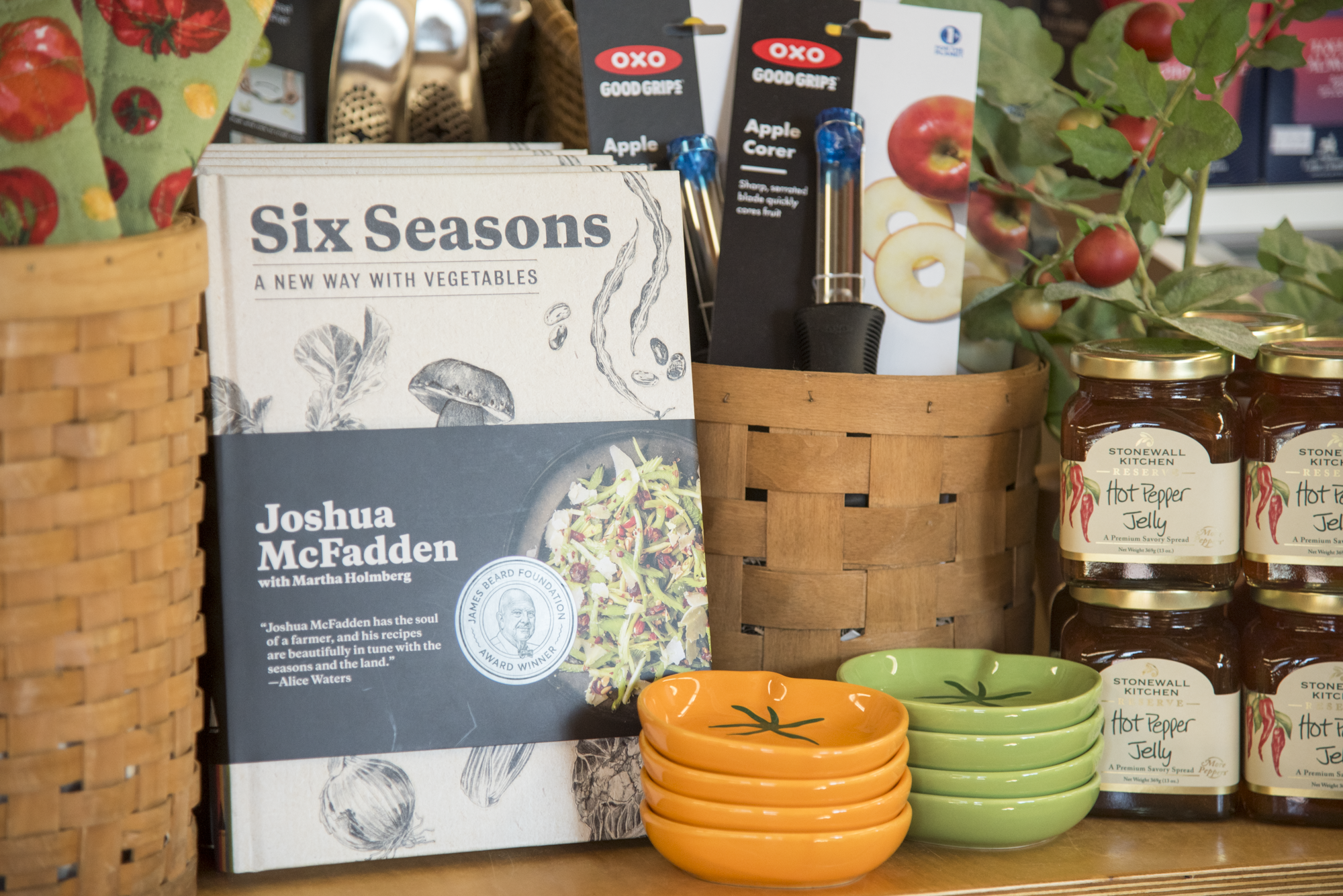 The cookbook 'Six Seasons: A New Way with Vegetables' sits on a shelf alongside kitchen gadgets, textiles, dishes, and pantry goods.