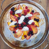 An RSVP Endurance Precision Pierced Pizza Pan sits on a wooden table. The pan has a fruit pizza on it.